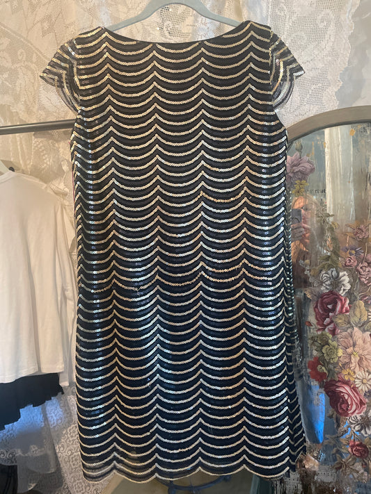 A sleeveless fit-and-flare dress with a shiny black base and a repeating metallic gold wavy scallop pattern, hanging on a hanger in a retail setting.