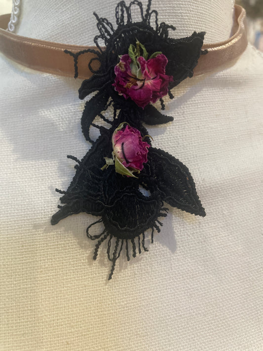 A close-up of a velvet and black lace necklace decorated with two dried pink rosebuds and black lace appliqués, displayed on a cream fabric mannequin bust.